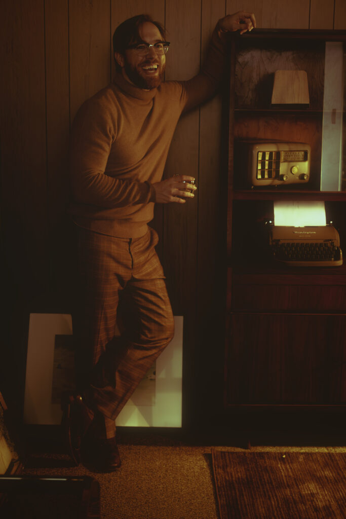 Rob smiles in a candid photo. He wears a camel-colored turtleneck and brown plaid pants and leans against a vintage bookshelf with an old radio and a typewriter. In his other hand he holds a cocktail glass.